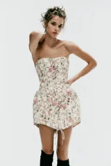Vestido corto strapless de corte corsetero, con estampado floral en tonos rosados y verdes sobre fondo blanco. Presenta detalles de tiras cruzadas ajustables en el delantero con lazada y varillas interiores.