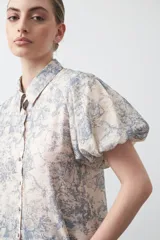 Camisa de manga corta abullonada, con cuello camisero y botones frontales. Presenta un estampado de inspiración barroca en tonos azul grisáceo sobre fondo beige claro.