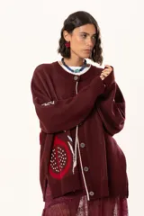 Saco de punto color vino con diseño holgado, cuello redondo y mangas largas. Presenta un gran bordado central en la espalda con forma de flor en tonos crema y rojo, acompañado de un pequeño bordado de sol y una frase bordada en la parte inferior.