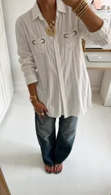 Chemise larga de rayas color blanco, con cuello camisero, manga larga y bolsillos frontales con detalles de argollas metálicas.