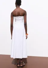 Vestido midi de corte evasé, color blanco, con escote palabra de honor y detalle fruncido en los laterales. Cuenta con cierre de cremallera en la parte posterior.
