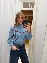 Blusa de denim fino color celeste con cuello alto y volados. Presenta detalles de ribetes en terciopelo rojo en el frente y hombros, mangas largas con puños abotonados y diseño romántico.