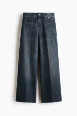 Jean tobillero de denim azul con leve elasticidad, tiro alto y perneras amplias tipo wide leg. Presenta bolsillos delanteros tipo parche con solapa y botones metálicos, además de pliegues marcados al frente y en la espalda.