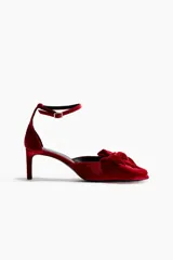 Zapatos de tacón tipo pump en terciopelo rojo, con punta fina y un lazo grande decorativo en el empeine. Presentan una pulsera ajustable al tobillo con cierre de hebilla y tacón fino forrado.