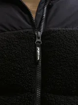 Campera puffer con cuello alto y cierre frontal, confeccionada en tejido teddy negro con paneles superiores lisos. Cuenta con bolsillos laterales y relleno de poliéster.
