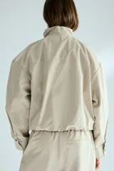 Campera corta de sarga de algodón, color beige, con cuello elevado ajustable por cordón, solapa rompevientos con botones a presión ocultos y cierre. Presenta bolsillos delanteros insertados, mangas ranglán con hombros caídos y bajo globo que genera una silueta abullonada.