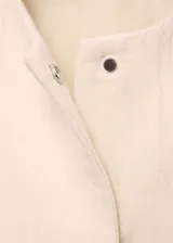 Chaqueta de algodón color crema, con cuello redondo, cierre frontal oculto y mangas largas con detalles de desflecado en los laterales.