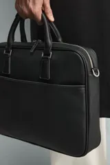 Maletín negro con textura grabada, diseño estructurado y asas de mano. Cuenta con cierre principal de cremallera, bolsillo frontal plano y bolsillo interior para dispositivos electrónicos de hasta 13 pulgadas. Incluye asa de hombro ajustable y extraíble.