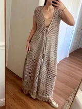 Chaleco largo sin mangas, tejido en punto calado tipo red con hilo de algodón y lurex, color marrón. Presenta escote profundo en V, tiras para atar en el frente, tajo trasero y flecos en el ruedo.