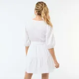 Vestido corto blanco con diseño de bordado inglés, escote en V con botones frontales, mangas abullonadas tres cuartos y detalle de lazo ajustable en la cintura con falda escalonada.