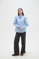 Camisa celeste de manga larga con cuello alto y volados, detalles de frunces en hombros y puños, y cierre frontal con botones.