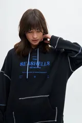 Canguro color crudo con capucha, cordón de ajuste celeste, bolsillo tipo canguro, puños y ruedo acanalados, y estampado frontal con la frase "The Seashells".