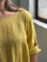 Blusa mostaza de lino, con cuello redondo, mangas tres cuartos con presilla y botón, y detalle de pliegues en la parte inferior.