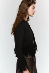 Chaqueta de punto color verde oscuro, con cuello redondo, manga larga y cierre frontal con un solo botón. Presenta detalles de flecos en el ruedo y en los puños.