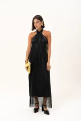 Vestido largo negro de cuello halter con diseño cruzado, confeccionado en tela con leve brillo. Presenta una silueta fluida y terminación con flecos en el ruedo.