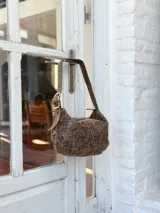 Cartera tipo hobo de cuero con acabado texturizado y efecto envejecido en tono marrón visón. Presenta una correa ajustable con hebilla metálica y cierre superior.
