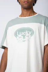 Remera de corte holgado con diseño color block, combinando paneles en color verde menta y blanco. Presenta un estampado frontal con la palabra 'Lucky' dentro de un óvalo y dibujos de flores con lentes de sol.