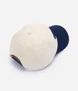 Gorra de béisbol con diseño bicolor, cuerpo en color crema y visera en azul marino. Presenta un bordado frontal con lettering en relieve color azul.