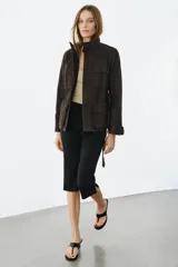 Chaqueta de estilo safari confeccionada en mezcla de algodón con acabado encerado. Presenta cuello alto con trabilla ajustable y hebilla metálica, cierre frontal con cremallera y botones ocultos bajo solapa, bolsillos de plastrón con solapa en el delantero y cinturón ajustable del mismo tejido para anudar en la cintura.