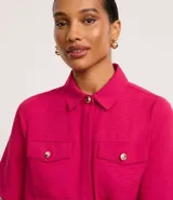 Camisa color fucsia de tejido airflow, con cuello camisero, mangas cortas con pliegues, cierre frontal con botones metalizados y bolsillos con solapa en el pecho.