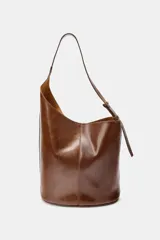 Bolso tipo saca de diseño asimétrico confeccionado en piel de color marrón. Cuenta con un asa de hombro ajustable y cierre mediante imán. Incluye una cartera con bolsillo extraíble en su interior.