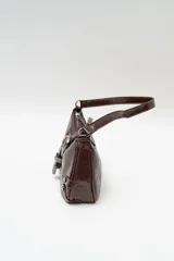 Bolso de hombro tipo baguette en color negro con acabado brillante. Presenta un diseño estructurado con una hebilla decorativa metálica en el frente y detalles de tachas plateadas en los laterales.