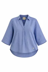 Blusa azul de lino con cuello camisero, escote en V y mangas tres cuartos con vuelta. Presenta un diseño holgado con dobladillo redondeado y un pequeño parche con logo bordado en el lateral inferior.