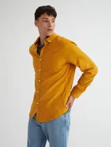 Camisa de manga larga color mostaza confeccionada en pana fina, con bolsillo en el pecho, botones de pasta, bordado en el ruedo y etiqueta de marca en la tapeta de los botones.