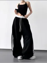 Pantalón deportivo negro de calce extra amplio (wide leg) con cintura elástica blanca y cordón ajustable blanco. Presenta tres tiras verticales blancas a lo largo de las costuras laterales.