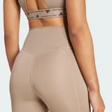 Calza deportiva de entrenamiento Adidas Optimé Essentials, color tostado de tiro alto con tecnología AEROREADY. Cuenta con cintura que se mantiene firme y costuras selladas.