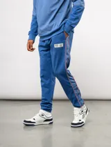 Pantalón deportivo de piqué color azul marino, con franjas laterales en contraste rojo y blanco. Presenta el escudo del Club Nacional de Football bordado en la pierna derecha y una etiqueta conmemorativa en la izquierda.