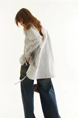 Camisa oversize de corte holgado con estampado de rayas verticales. Presenta cuello camisero, cierre frontal con botones a contraste y cordones integrados en la cintura para ajustar la silueta. Posee mangas largas con puños rectos.