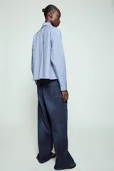 Camisa de algodón color celeste, con cuello alto y lazo integrado en el mismo tejido. Presenta cierre frontal con botones, manga larga con puños y un bolsillo frontal en el pecho. Diseño de corte recto.