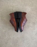 Zapato acordonado de cuero vacuno color marrón con suela de goma negra y cordones marrones.