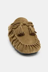 Mocasín destalonado tipo mule confeccionado en serraje color tostado, con detalle de fruncido y borlón en la parte delantera. Interior forrado en efecto borreguito y suela plana.