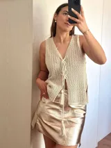 Chaleco tejido en punto calado con hilo de algodón y lurex, de color negro con destellos plateados. Presenta escote profundo en V, cierre frontal con cordones entrelazados terminados en borlas y un corte recto y holgado.