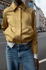 Chaqueta bomber de efecto ante en color tostado, con cuello solapa, manga larga y bolsillos laterales. Presenta un diseño con bajo acabado en pliegues que aporta volumen y cierre frontal con cremallera y botones a presión.