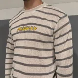 Sweater tejido a rayas horizontales grises y blancas con logo amarillo bordado en el pecho.