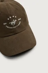 Gorra de seis paneles de color marrón oscuro, confeccionada en tejido de algodón. Cuenta con un diseño bordado en la parte frontal que incluye el texto "TEXAS OUTLAWS", la fecha "1999" y una silueta con estrellas.