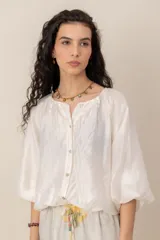 Blusa blanca de diseño amplio, con mangas abullonadas, escote redondo fruncido y cierre frontal de botones nacarados. Cuenta con elástico en la cintura que genera efecto blusón.