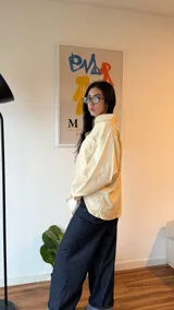 Camisa oversize de corte amplio con estampado de rayas verticales finas en tonos amarillo y azul. Presenta cuello clásico, cierre frontal con botones, mangas largas con puños abotonados y un bolsillo en el pecho.