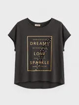 Remera de algodón con corte relajado y diseño asimétrico, con espalda más larga que el frente. Presenta un estampado frontal rectangular con marco dorado, texto motivacional y una pluma decorada con pequeñas perlas.