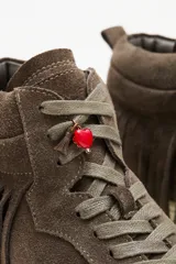 Zapatillas de caña media confeccionadas en cuero vacuno descarne color marrón. Presentan un diseño con flecos laterales, cordones al tono y un pequeño dije de corazón rojo en la parte superior de la lazada. Cuentan con suela de goma en color crema.