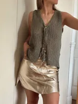 Chaleco tejido en punto calado con hilo de algodón y lurex, de color negro con destellos plateados. Presenta escote profundo en V, cierre frontal con cordones entrelazados terminados en borlas y un corte recto y holgado.