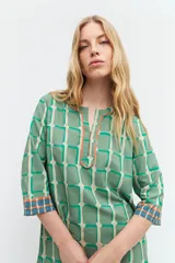 Túnica verde con estampa geométrica de cuadrados en verde más oscuro y blanco. Tiene escote en V con ribete marrón y mangas tres cuartos con ribete geométrico en puños y ruedo.