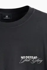 Remera negra de algodón con cuello redondo acanalado y mangas cortas de hombros caídos. Presenta un pequeño estampado tipográfico en el pecho con las palabras "No Defeat Just Glory".