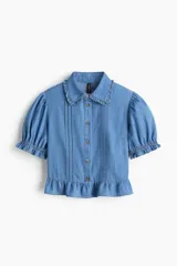 Blusa corta confeccionada en denim de algodón, con diseño entallado, cuello con volados, mangas cortas abullonadas con puños elásticos y botonadura frontal. Presenta alforzas decorativas en el frente y un volado en el ruedo.