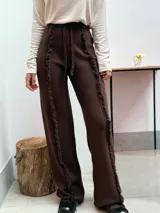 Pantalón de tejido de punto color marrón, de corte recto y holgado. Presenta detalles de costuras con flecos verticales a lo largo de las piernas y cintura elástica con cordón de ajuste.