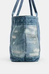 Bolso tipo shopper de hombro efecto denim, confeccionado con paneles de mezclilla de diferentes lavados y detalles deshilachados. Cuenta con doble asa de hombro y bolsillo interior con cremallera.