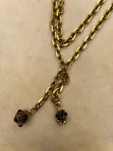 Collar de cadena de eslabones gruesos en tono dorado, con diseño tipo lazo que termina en dos dijes colgantes: uno esférico con detalles labrados y piedras negras, y otro con forma de flor decorado con cristales negros.
